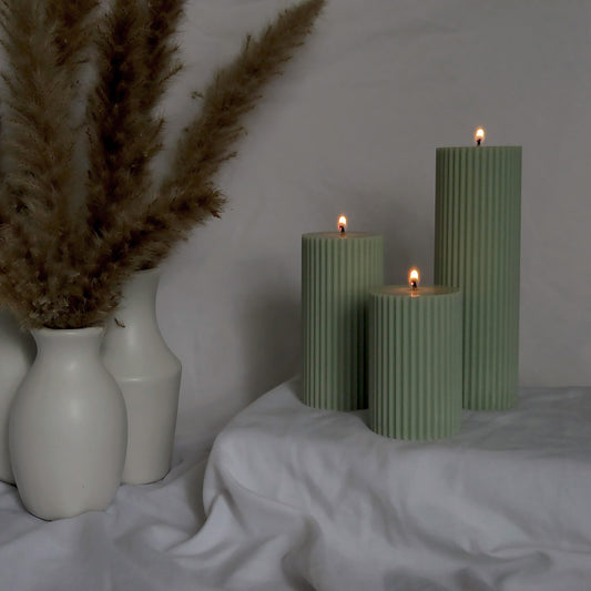 Ribbed sage pillar candles displayed decoratively, showing the clear size differences between the 3 options. The candles are lit, demonstrating the flame size. Available as scented or unscented versions, the ribbed effect of the soy pillar candles adds a unique texture, that adds a sophisticated touch to any home decor style.